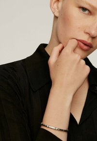 Camisa negra de cuello con textura vertical; mano reposando en el mentón; pulsera plateada en la muñeca; maquillaje natural y tonos neutros.