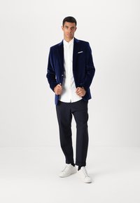 Navy fluwelen blazer met een enkele knoop, een witte overhemd eronder, navy broek en witte sneakers. Zachte textuur, getailleerde pasvorm.