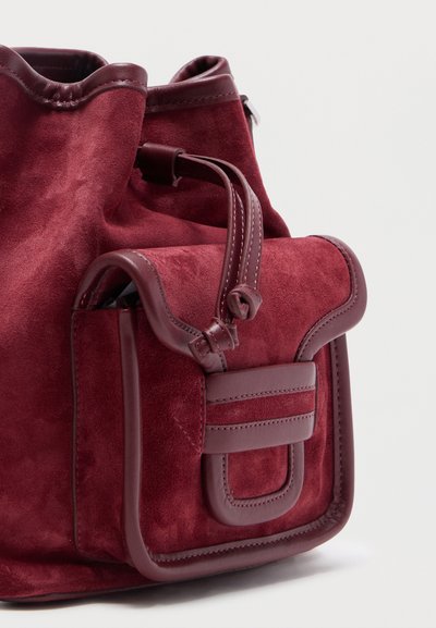 Σακίδιο πλάτης από σουέτ σε βαθύ βουργουνδί χρώμα με δερμάτινες λεπτομέρειες, που διαθέτει μπροστινή θήκη με καπάκι ασφαλισμένη με δερμάτινο λουρί και ραφές για διακοσμητικό αποτέλεσμα.