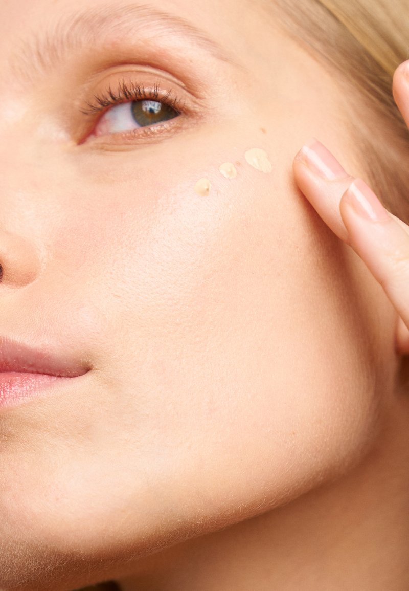 Liquid foundation in a light beige shade is applied to the skin. The texture appears smooth and dewy on a lightly textured complexion.