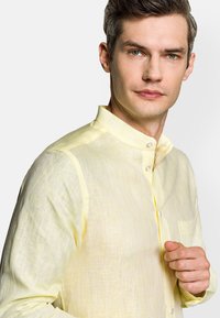 Jasnożółta lniana koszula z kołnierzykiem stojącym, subtelną fakturą i zapięciem na guziki z przodu. Posiada kieszeń na piersi oraz dopasowany krój.