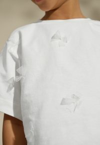 Biała bawełniana koszula z teksturowanej tkaniny z małymi, przezroczystymi akcentami w postaci kokardek. Krótkie rękawy i luźny krój dla wygody. Minimalistyczny design.
