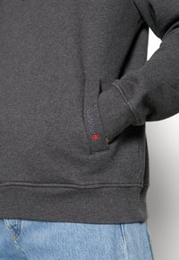 Ciemnoszara bluza z kapturem z kieszenią z przodu, na której znajduje się drobna haftowana logo „Barbour”, w zestawieniu z jasnoniebieskimi dżinsami.