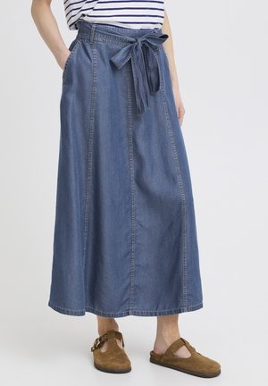 Žena v modrej rifľovej maxi sukni so zaviazaným opaskom, pruhovanom topu a hnedých semišových nazúvacích topánkach so sponami.