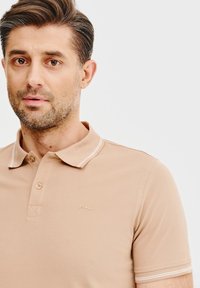 Beżowa koszulka polo z teksturowanej tkaniny, z dwoma guzikami przy zapięciu, kontrastowym wykończeniem kołnierzyka oraz małym złotym logiem na piersi.