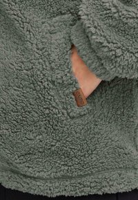 Fuzzy, grøn fleece-pullover med høj krave, sidelomme og læderlogo-patch med teksten "OXMO." Struktureret og blød stof.