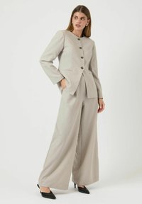 Ljusgrå skräddarsydd kostym med en figursydd, knäppt blazer och vida byxor. Slätt tyg med subtil textur och svarta klackskor.