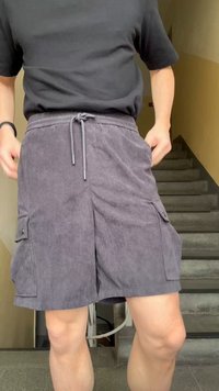 Άτομο που φορά σκούρα γκρι βερμούδα κοτλέ cargo και μαύρο πουκάμισο με κοντό μανίκι, στέκεται κοντά σε μια εσωτερική σκάλα.