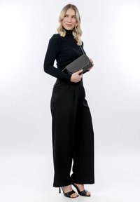 Suéter negro de cuello alto con textura acanalada, pantalones negros de pierna ancha y elegantes tacones altos negros. Sosteniendo un bolso de mano negro con un detalle plateado y cremallera.