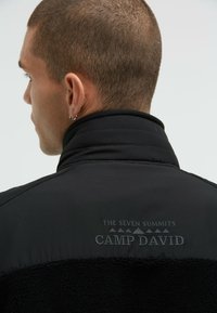 Black jacket with a padded collar and soft fleece texture, featuring "THE SEVEN SUMMITS CAMP DAVID" text embroidered on the back.