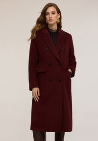 Cappotto in lana borgogna con design a doppiopetto, bottoni neri, rever a doppio taglio, tasche laterali e vestibilità su misura, indossato sopra un top testurizzato.