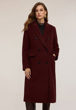 Cappotto in lana borgogna con design a doppiopetto, bottoni neri, rever a doppio taglio, tasche laterali e vestibilità su misura, indossato sopra un top testurizzato.