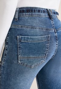 Gros plan sur une personne portant un jean en denim bleu avec des coutures visibles, une poche arrière, et un haut blanc rentré à l'intérieur.