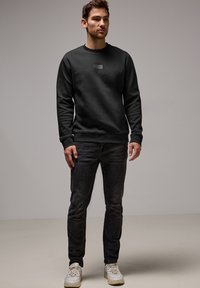 Sort sweatshirt med tekstureret logo, rund hals og lange ærmer, parret med mørke jeans og hvide sneakers på en neutral baggrund.