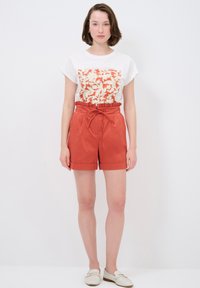 T-shirt bianco con stampa floreale rossa, abbinata a pantaloni shorts a vita alta color ruggine, con coulisse e orli arrotolati. Scarpe slip-on color avorio.