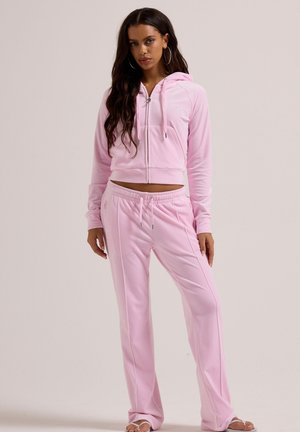 Frau, die steht, trägt einen hellrosa Kapuzenpullover mit Reißverschluss und passende Hose mit Kordelzug, Ohrreifen und Sandalen, neutraler Hintergrund.