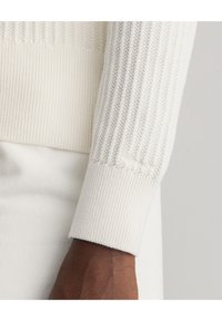 Bílý žebrovaný pletený rukáv se strukturovaným vzorem, měkký materiál a kontrastní hladké pletení na základně.