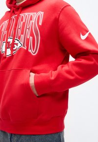 Άτομο που φοράει κόκκινο φούτερ με κουκούλα των Kansas City Chiefs με λογότυπο Nike στο μανίκι, με το χέρι στην μπροστινή τσέπη και ορατό μπλε τζιν