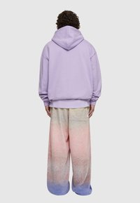 Lila, oversized kapucnis pulóver, állítható kapucnival, széles lábú nadrággal párosítva, amelyen lágy rózsaszín és kék árnyalatú gradiens minta található.