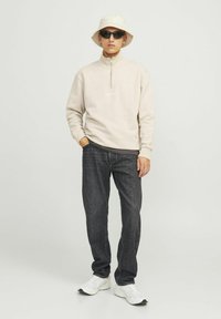 Beige ritsvest met een kraag, donkere spijkerbroek, witte sneakers en een crèmekleurige bucket hat, gecombineerd met zwarte zonnebril.