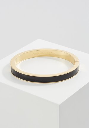 Złota bransoletka typu bangle z czarną emalią, leżąca na białej powierzchni, z grawerem wewnątrz: „MIEJ ASA W RĘKAWIE.”