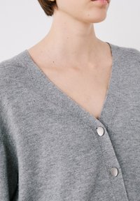 Cardigan grigio realizzato in materiale morbido con scollatura a V, dotato di due bottoni metallici e un motivo a maglia texturizzato.