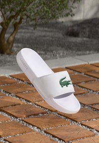 Sandale blanche à glissière avec une large sangle arborant un logo de crocodile vert. Fabriquée en matériau synthétique au toucher lisse. Design léger.