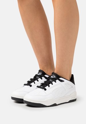 White low-top sneakers with black laces and accents, featuring a smooth leather exterior, textured heel, and perforated toe area.