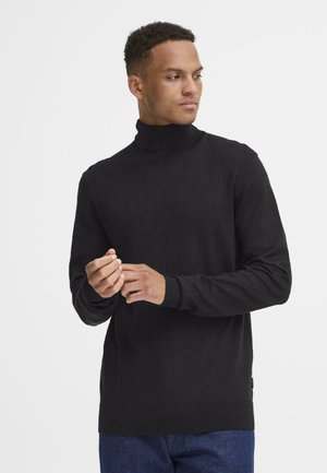 Jersey de cuello alto negro con detalles de canalé en el cuello y los puños. Textura suave, diseño ajustado. Llevado con jeans azules.
