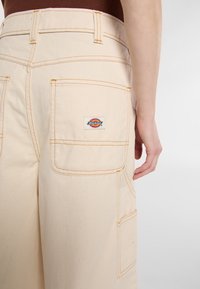 Pantaloni da lavoro beige con cuciture arancioni, multiple tasche e un'etichetta con il logo Dickies visibile sul retro. Tessuto in canvas di cotone.