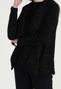 Černý texturovaný kardigan s volným střihem, zdobený tečkovaným leskem, s dlouhými rukávy a otevřeným předním dílem, nošený přes tmavé kalhoty.