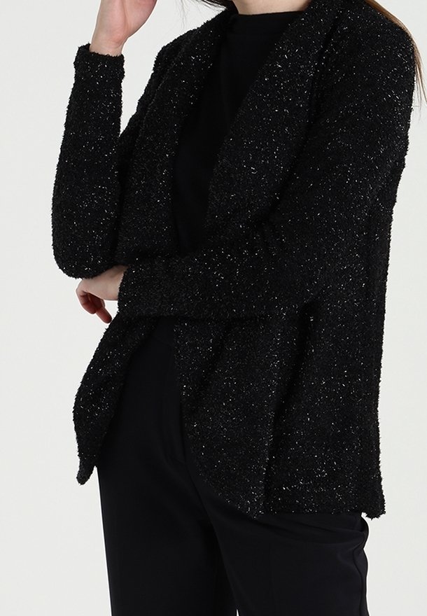 Černý texturovaný kardigan s volným střihem, zdobený tečkovaným leskem, s dlouhými rukávy a otevřeným předním dílem, nošený přes tmavé kalhoty.