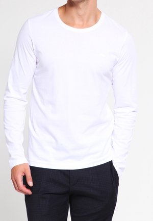 Camiseta blanca de manga larga con cuello redondo y textura suave, combinada con pantalones oscuros de estampado sutil. Branding discreto en el pecho.