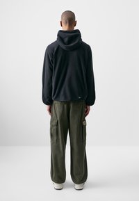 Sweatshirt preto com capuz e um acabamento texturizado, combinado com calças cargo verde-oliva. Inclui bolsos laterais e um corte relaxado. Detalhe do logótipo em branco visível.