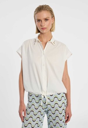 Blonde Frau trägt ein lockeres weißes kurzärmeliges Hemd mit verziertem Kragen und gemusterte, wellenförmige gestreifte Hose, steht vor weißem Hintergrund.