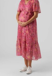 Robe de maternité rose à fleurs avec des manches courtes, une taille à nouer et un ourlet en volants, associée à des baskets blanches. Tissu semi-transparent.