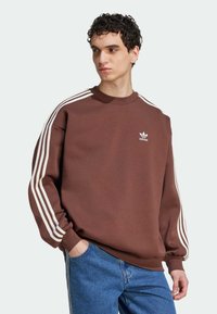 Smeđa dukserica s bijelim trostrukim uzorkom na rukavima, okrugli izrez, mekana tekstura i mali bijeli Adidas logo na prsima.