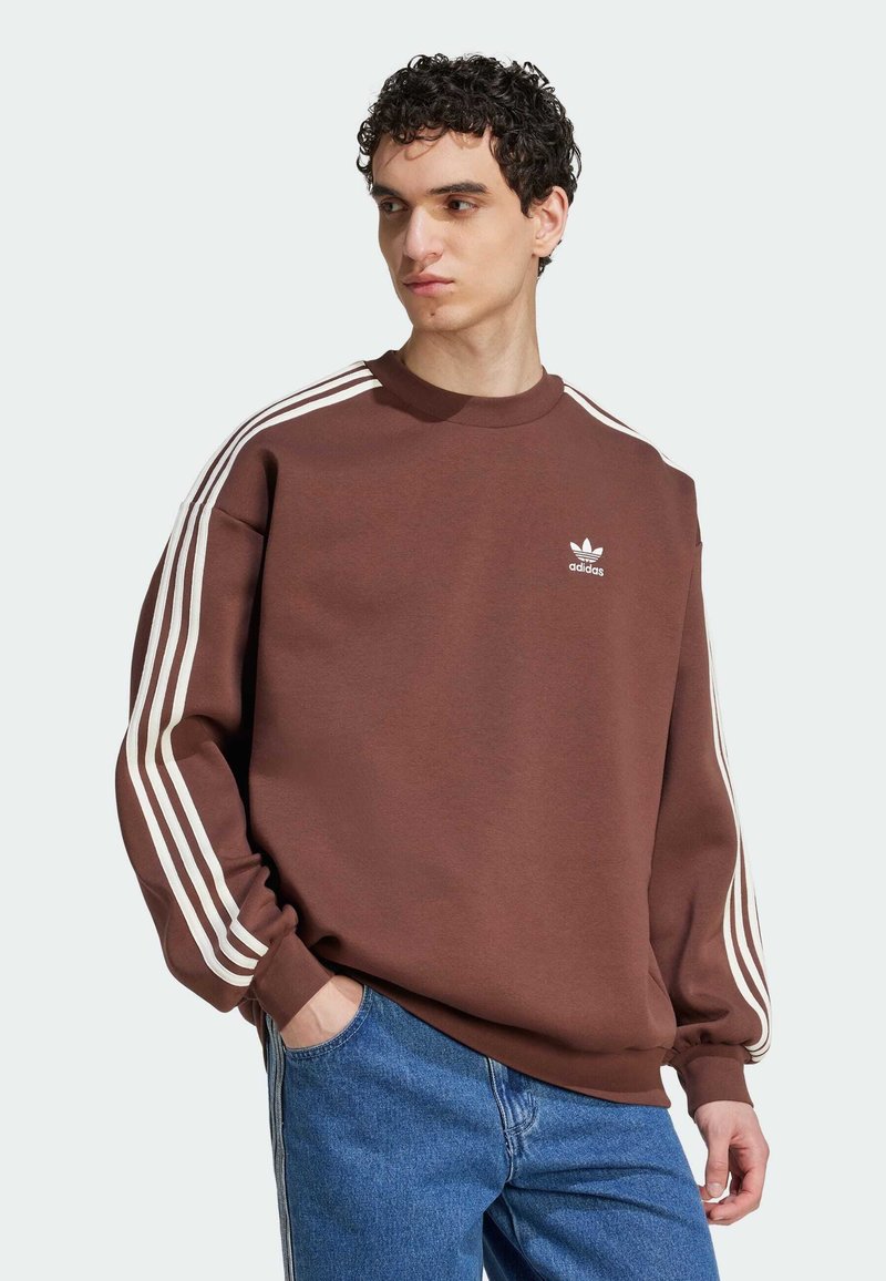Smeđa dukserica s bijelim trostrukim uzorkom na rukavima, okrugli izrez, mekana tekstura i mali bijeli Adidas logo na prsima.