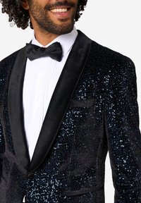 Čierny smokingský sak s flitrami a saténovými klopami, ktorý predvádza štruktúrovaný povrch a ostrý siluetu, nosený nad bielou skladanou košeľou a motýlikom.