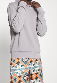 Sudadera gris con cuello alto y mangas largas, combinada con pantalones cortos estampados que presentan diseños geométricos en naranja, negro y turquesa.