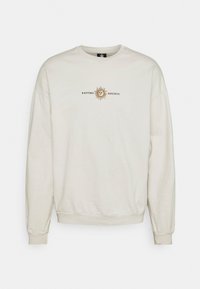 Felpa beige chiaro con scollo rotondo, polsini e orlo a costine, con una grafica di un sole stampata e il testo "KAOTIKO SOCIETY."