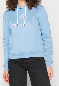 Lichtblauwe hoodie gemaakt van zacht materiaal. Het heeft een zak aan de voorkant, witte trekkoorden en roze logotekst met een diamantaccent.