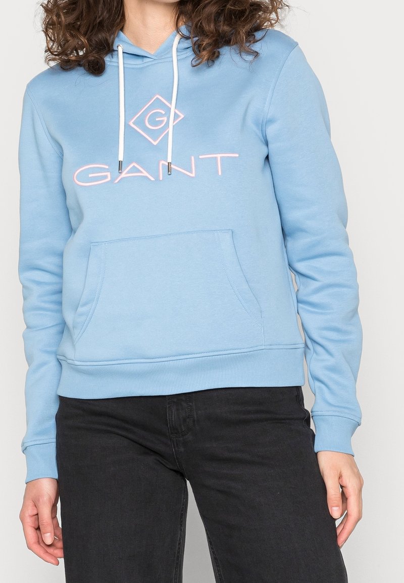 Lichtblauwe hoodie gemaakt van zacht materiaal. Het heeft een zak aan de voorkant, witte trekkoorden en roze logotekst met een diamantaccent.