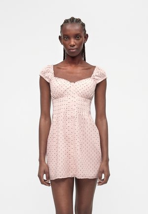 Kobieta z warkoczami ubrana w jasnoróżową, krótką sukienkę zapinaną na guziki z czarnymi grochami, stojąca na jednolitym tle.