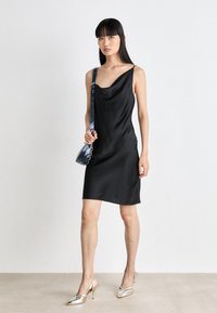 Black satin slip dress with a cowl neckline and thin straps, paired with silver heeled shoes. Includes a blue patterned shoulder bag.