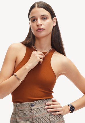 Débardeur marron sans manches en tissu texturé, porté avec un pantalon taille haute à carreaux. Les accessoires comprennent un collier en chaîne argenté et un bracelet.