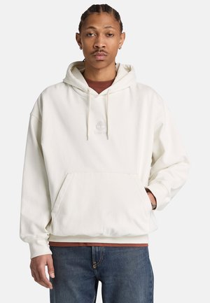 Jonge man met gevlochten haar draagt een witte hoodie over een bruin overhemd en blauwe spijkerbroek, staat met één hand in de zak.