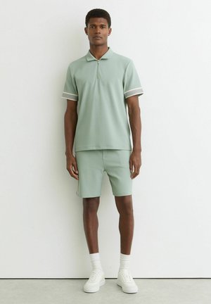 Mężczyzna w jasnozielonej, krótkim rękawem, zapinanej na zamek koszulce polo i dopasowanych szortach, w białych tenisówkach i skarpetkach, na tle białej ściany.