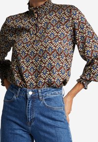 Braun gemusterte Bluse mit blauen und gelben Akzenten, ausgestattet mit einem hohen Rüschensaumkragen und Rüschenmanschetten, getragen mit blauen High-Waist-Jeans.