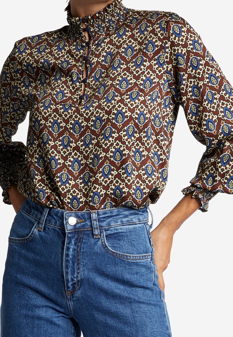 Braun gemusterte Bluse mit blauen und gelben Akzenten, ausgestattet mit einem hohen Rüschensaumkragen und Rüschenmanschetten, getragen mit blauen High-Waist-Jeans.